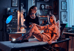 young tattoo master is standing crossed her hands while attractive girl is waiting her couch
